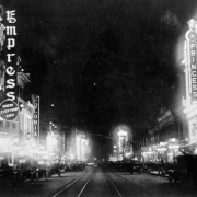 Nighttime view of theaters along Curtis Street in downtown Denver, Colorado. Automobiles line the street. Signs read: "Empress Where Everybody Goes," "Colonial," "Mozart," "Royal," "Drugs," "Iris," "The Nanking," "Pool," "Hotel," "Princess," and "Onyx."