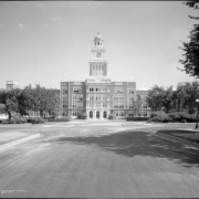 View of west facade entry of East High School from City Park Esplanade and Sixteenth Street, Denver, Colorado; brick and terra cotta building with clock tower; shows man seated on front steps under arched entry, trees, hedges and flowers, concrete curbs, and streetlights along streets.