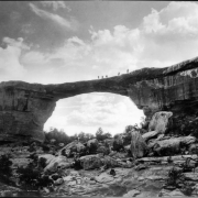 Owachomo Natural Bridge, Armstrong Canyon, Natural Bridges National Monument, San Juan County, near Blanding, Utah. People pose on top of the sandstone formation also known as Little Bridge, with pinyon and  juniper trees on slope below.