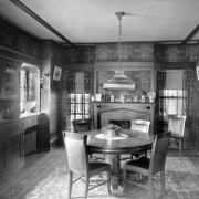 Interior view of a Denver, Colorado, residence; dining room furnishings include a brick fireplace, wood paneling, molding, Art Nouveau wallpaper, a pedestal table, leather upholstered wood chairs, Oriental rug,  stained glass lamp, built-in buffet, crystal punch bowl, silver coffee pot, and china cabinet. Vases and photographs top the mantle.