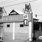 View of the Crystal Grill at 2631 Larimer Street in the Five Points neighborhood of Denver, Colorado. Shows a single-story storefront addition to a private residence. The twentieth-century brick addition has three small windows with brick sills, a single door entrance, and a parapet. A sign running across the facade reads: "Crystal Grill." Other signs on the building advertise 7-Up and Schlitz.