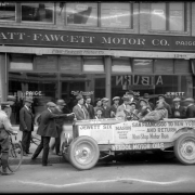 Crowd gathers around automobile crew who just completed an "Automobile Endurance Run" parked in front of the Platt-Fawcett Motor Co., 1249-1255 Broadway, Denver, Colorado; car is a Jewett Six model made by Paige, with lettering on side: "San Francisco to New York and Return, Non- Stop Motor Run, Mason Safety Cord Tires, Veedol Motor Oils;" town names of Reno and Marysville, Calif. are written on car; man with bicycle at left.