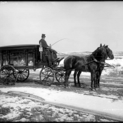 View of a horse-drawn hearse on a muddy road, probably in Denver, Colorado; the driver wears a top hat and coat. Snow covers fields.