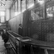 Interior view of the Daniels and Fisher store, in Denver, Colorado; shows glass counters and display cases. A boy draws curtains by shelves of drawers.