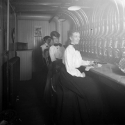 Interior view of a Daniels and Fisher store in Denver, Colorado; shows women seated at a pneumatic messaging device; one holds a rubber stamp by a "Staats Money Changer" machine.