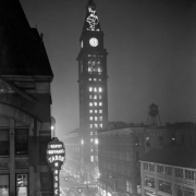 Night view of the Daniels and Fisher Stores tower, in Denver, Colorado; shows 16th (Sixteenth) Street with Christmas decorations, and neon signs: "Harry Huffman's Tabor Musical," and "D & F (Santa Claus)."