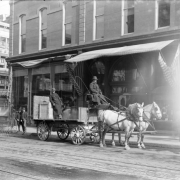 View of a Daniels and Fisher store delivery wagon and horses in Denver, Colorado; shows men and a teenaged boy in bowler hats. Storefronts are in the background.