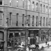 View of the Daniels and Fisher store in Denver, Colorado; shows glass storefront windows, exterior sconce lamps, and a horse and buggy.