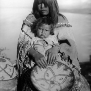 Seated outdoor portrait of a Native American (Apache) young woman and child. The woman wears a painted and fringed buckskin poncho over a print dress. She holds the small girl on her lap, her hands are placed in front of the child and hold a woven tray basket.