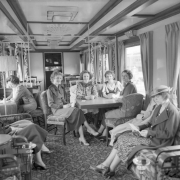 Women in high heels, dresses, skirts and hats pose on furniture inside a Denver and Rio Grande Western Railroad dining lounge car, refurbished from a Western Pacific dining car, parked possibly in the Denver, Colorado railroad yards. Some of the women play cards. Interior design is Art Deco with arm chairs, sofas, tables, lamps, smoking stands and ornate metal wall partitions. Upholstery features floral and geometric motifs. Possibly taken during the Summer of 1939.