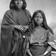 Studio portrait (sitting and standing) of Native American (Kiowa) women; one woman is Big Tree's sister. The women wear dresses, one wears a fringed shawl.