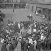 Taos Pueblo Indians and tourists form a circle around dancers with a United States flag in the Taos Plaza, Taos, New Mexico. Native Americans (Taos Pueblo) are wrapped in blankets, the sightseers wear hats. Onlookers stand on porches or portals of enclosing buildings. A sign reads: "Post Card Views, Indian Curios, Kodak Finishing."