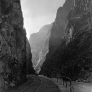View along the Arkansas River and the Denver and Rio Grande Western railroad tracks, with the Hanging Bridge in distance, Royal Gorge, Fremont County, Colorado. Wooden Canon City water supply pipeline shows tunneling in and along the steep granite wall along river.