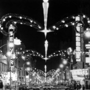 Nighttime view of 16th (Sixteenth) Street in downtown Denver, Colorado. Automobiles drive under Christmas lights and decorations that span the street. Signs read: "Denver," "Baurs," "Douglas Optical," and "Paramount."