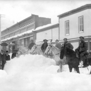 View of a heavy snowfall on Sixth (6th, Alpine) Street in Georgetown, Colorado. The Spruance Building and Kneisel-Curtis-Seifeld building are on the south side of the street. Five men ride large horses; five men stand (wearing heavy coats and hats) surrounded by piles of snow.