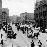 African American men march in probably a protest parade with horse drawn wagons, farm implements and wheeled carts on Seventeenth Street, in Denver, Colorado; shows the Johnson Building, the Denver Club, the Savoy Hotel, pedestrians, automobiles, and Sign reads: "New - Times," and has a barometer.