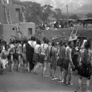 View of Native American (Taos Pueblo) men at Taos Pueblo, New Mexico, wearing moccasins, breechcloths, and body paint, during a festival. They carry branches; adobe pueblo dwellings and white spectators are in the background.