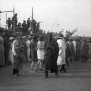 View of the San Geronimo festival at Taos Pueblo, New Mexico; shows Native American (Taos Pueblo) men wearing body paint and holding branches. White men and women watch from the side.