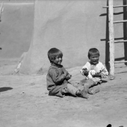 Portrait of Native American (Taos Pueblo) boys by a ladder and adobe walls at Taos Pueblo, New Mexico; they play with a puppy.