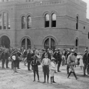 Colorado National Guard, mustered to combat the labor strike of the Western Federation of Miners, march on Curtis Street in front of the brick Armory building at 2565 Curtis, Denver, Colorado. A crowd, including two Black children, stand and watch.