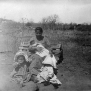 Annie Colliere, a Native American (Plains) woman, poses outdoors with three children. She holds a baby wrapped in a blanket; one of the children wears a bonnet; the other has her head covered by the baby's blanket. A brush shelter and trees are in the distance.