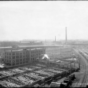View of Denver Union Stock Yards Company cattle pens and construction of Livestock Exchange building in Denver, Colorado; also shows railroad tracks and cars, corrals of sheep, and the Omaha and Grant Smelter smokestack.