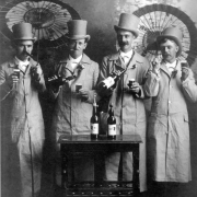 Studio portrait of Charlie Nelson, Thomas C. Browne, Douglas Patilla, and Charles Smith in Denver, Colorado. The men hold parasols and pour beer from bottles. They wear top hats and overcoats.