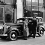 Men pose near an automobile in Denver, Colorado. They wear overcoats and hats. Lettering on a nearby window reads: "Pontiac."