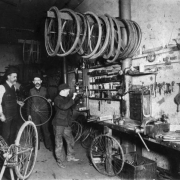 Interior view of a bicycle repair shop in Denver, Colorado; shows men, a boy, a vise, workbench, drill press, bicycle parts, and tools.