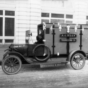 A man sits in the driver's seat of a delivery truck in Denver, Colorado. The rear of the truck is designed to look like a trunk. Signs on the truck read: "A. E. Meek Trunk & Bag Co." and "Trunks & Bags."