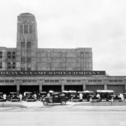 Men pose by pick-up trucks near the Blayney Murphy Company at 48th (Forty-eighth) Avenue and Gilpin Street (Brighton Boulevard) in the Swansea neighborhood of Denver, Colorado. The slaughterhouse has a tower topped with a flagpole. Lettering on the side of one of the trucks reads: "Mayflower Ham Blayney Murphy Company Service Car."