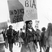 Native American men and women walk a picket line to protest perceived discrimination by the Bureau of Indian Affairs. Signs read: "Indins [sic] are red, B. I. A. what are you," "Stop persecuting Indians," and, "Give us BIA."