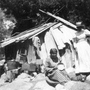 Two Native American (Miwok) women pose near a dilapidated wooden shack in Yosemite National Park, California. One woman, wearing a striped dress, sits holding a basket she is weaving. The other woman stands behind, wearing a long dress with leg o' mutton sleeves and a hat. The shack is loosely constructed and is surrounded by debris. A small bear is on the ground between the women.