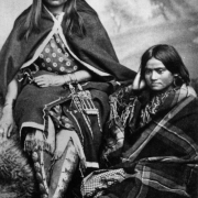 Studio portrait of Chi-wek-kiethe and her sister, Looking-For-Something-Good (Cha-wa-ke) sitting on a buffalo robe. The Native American Comanche women wear dresses wrapped with blankets, bracelets, a beaded bag, moccasins and leggings decorated with metal disks and long loose hair.