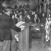 Captain Willaim S. Fairchild stands behind a wooden lectern addressing Japanese internees at Camp Amache, Prowers County, southeastern Colorado. Fairchild wears a military uniform; a United States flag stands near him. The front two rows of metal folding chairs are empty while several men stand against the back wall. Bare electric bulbs illuminate the block; fire extinguishers hang on support posts with angle braces.