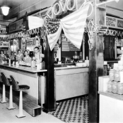 Interior view of a lunch counter at the Home Public Market, in Denver, Colorado; shows a man, women including Ruth Handler [third from right], a neon sign, tile floor, a United States flag, and signs: "Coca Cola," "Watermelon on Ice," "Beer On Draught," and "Greenwald's Soda Fountain."