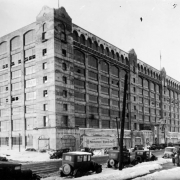 View of the newly constructed Montgomery Ward building at 501 South Broadway in the Baker neighborhood of Denver, Colorado. Automobiles and pedestrians are nearby. A sign near the building reads: "The New Rocky Mountain Home of Montgomery Ward & Co. The World's First Mail Order House Est. 1872 To Better Serve the People of Denver and the Mountain States."