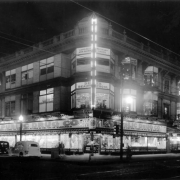 Nighttime view of the Enterprise Building on the corner of 15th (Fifteenth) and Champa Streets in downtown Denver, Colorado. Automobiles and pedestrians are near the building. Signs read: "Cut Rate Liquors and Wines," "The Purdy Studio," "Harco," "Geo. W. Fraser Tailor," "Katherine K. Corsets," "California Cotton Mills Co.," "Republic Drug Co.," "Denver Beauty School," "Coopers Flowers," and "Welcome V. F. W."