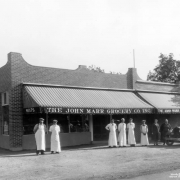 Employees pose outdoors near the John Marr Grocery Company in Denver, Colorado. The men wear aprons and caps. Lettering on the building's awnings read: "Mets," "The John Marr Grocery Co. Inc."