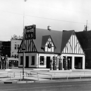 View of Frank L. Hansen's gas station at Broadway and Maple Avenue in the Baker neighborhood of Denver, Colorado. Fuel pumps and a two-car garage are near the building. A clock is above the main entryway. An electric sign reads: "Green Far-Go Gas."