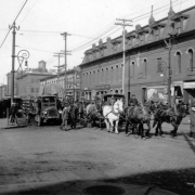 A team of horses pull a wagon with a large canvas covered object on Blake Street at 15th (Fifteenth) Street in downtown Denver, Colorado. Men walk nearby. Signs on buildings read: "The N. J. O'Fallon Supply Co.," "Goldberg's Harness Shop," "Soft Drinks," and "Cigars." A pick-up truck is beside the wagon.