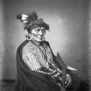 Seated studio portrait of a Native American man and Winnebago chief wearing a turban with roach and feathers, striped shirt, earrings and wrapped in a blanket. He holds a war club.