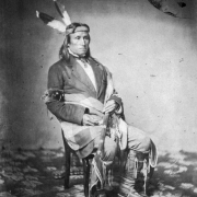 Seated studio portrait of  A-ki-pa (The Meeter), Native American man and warrior of the Sisiton Sioux, wearing a headband with two feathers, jacket, shirt and necktie, animal fur armband, leggings and a blanket around his waist; he hold a staff or calumet.