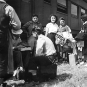 Japanese evacuees stand or sit with their suitcases and belongings in front of a Santa Fe and Topeka passenger train car. The men and women wait for the bus ride to Camp Amache, Granada Relocation Center, southeastern Colorado.