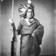 Seated studio portrait of He-kha-ka Ma-za (The Iron Elk), Native American man and Mdewakanton Sioux warrior, wearing a roach and feathers, jacket with shirt and necktie, and a blanket around his waist; he holds a ceremonial staff or spear.