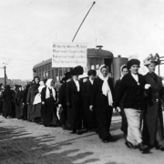 Women march in support of UMW coal miners on strike against CF&I in Trinidad, Las Animas County, Colorado; they carry a sign: "All Worthy Women My Sisters Have Ever Found It Thus If We Are True Friends To Others They'll Be True Friends To Us." A Trinidad Electric Railway trolley is by the parade; lettered brick building reads: "Trinidad Carriage Mfg. Co."