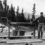 A man stands near a crank well hoist. Two buckets are attached to the rope.