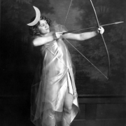 Helen Wilcox, a cast member in a production of "The Sunbird," poses indoors in Denver, Colorado. She holds a bow and arrow and wears a costume that includes sandals, cloak, and a headband with a moon.