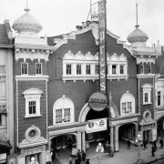 Rooftop view of the Empress Theater at 1615 Curtis Street in downtown Denver, Colorado. Pedestrians walk nearby. The three-story building has decorative trim and is topped with minarets. Signs on the building read: "Pantage's Vaudville" and "Now Showing 10 Acts Spring Festival Show."