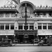 View of the Isis Theater at 1722 Curtis Street in downtown Denver, Colorado. The three-story building has an ornate theater facade with electric lights and signs that read: "First Run Conway Tearle and Faire Binney in" and "A Wide Open Space." Automobiles and a delivery truck that reads: "The Buerger Bros. Supply Co. Barbers Supplies."